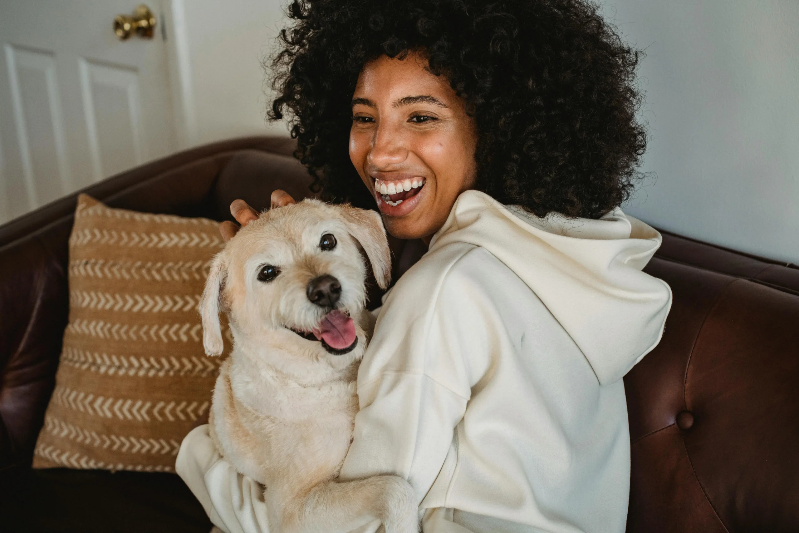 A smiling adult sitting on a couch holding a small dog inside a home